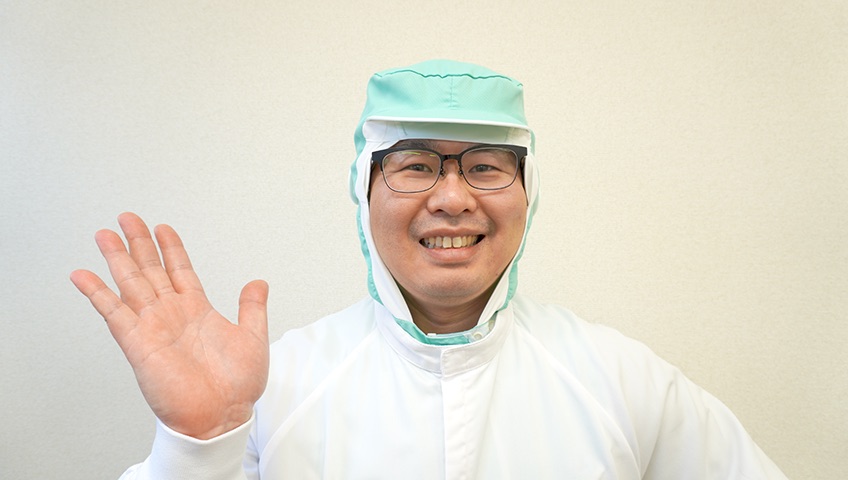 白い衛生衣と緑の衛生帽を着けて手を挙げ、笑顔で迎える工場長