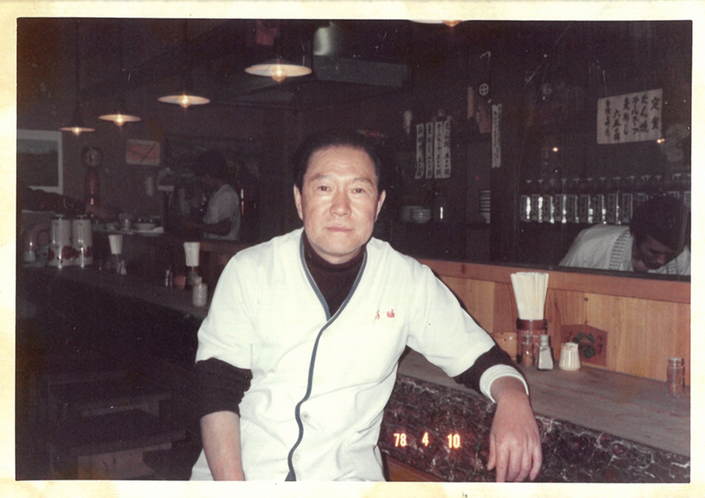 1978年4月、店のカウンター席に腰かける白い調理衣姿の創業者・大川原要
