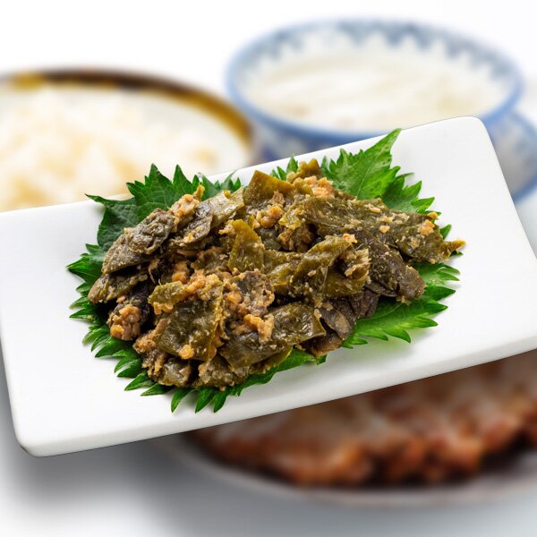 白い皿に大葉を敷いて盛り付けたみそなんばん。奥に麦飯の茶碗がぼかして写っている