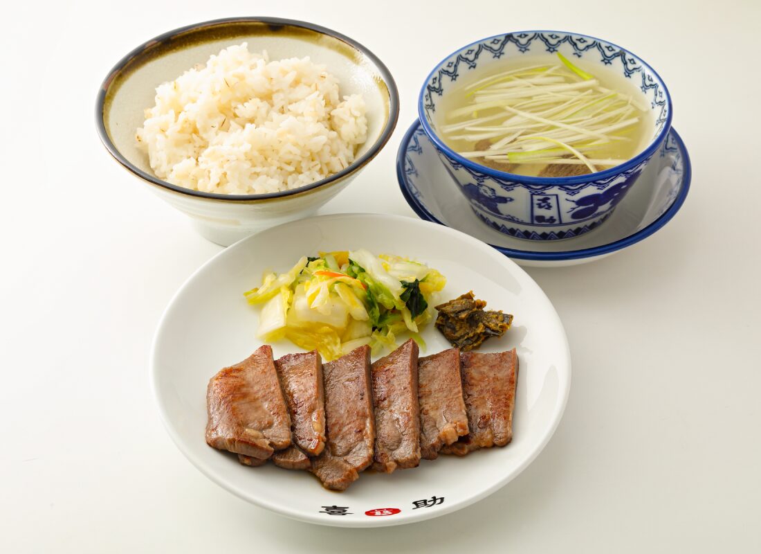 喜助の牛たん定食一式。喜助の銘が入った皿に牛たん焼きとお新香とみそ南蛮、麦飯の茶碗、白髪ねぎを浮かべた染付の碗のテールスープが並んでいる